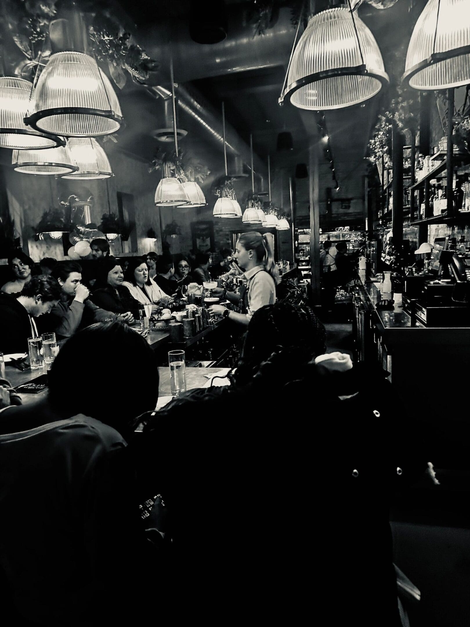 Black and white photo of a busy bar with a bartender serving customers under hanging lamps.