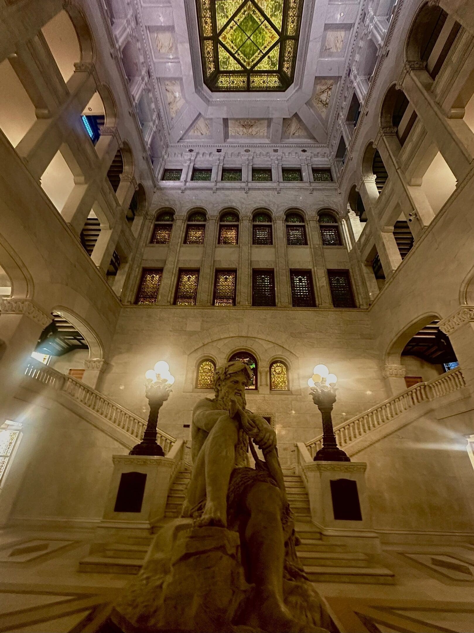 Statue in a grand hall with ornate stained glass ceiling and lamps.