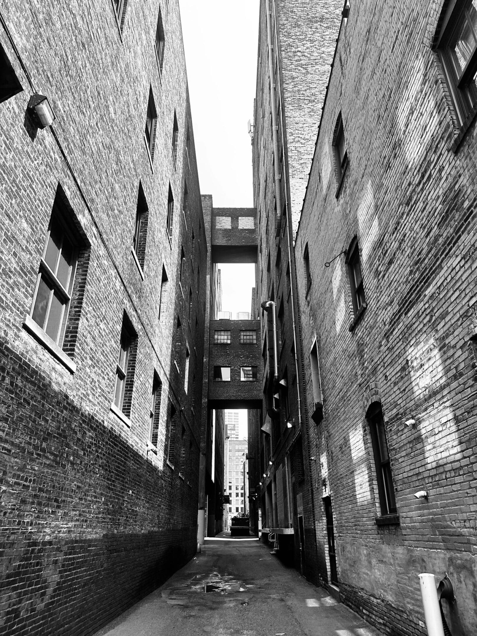 Narrow urban alley with brick buildings and sky bridges in black and white.