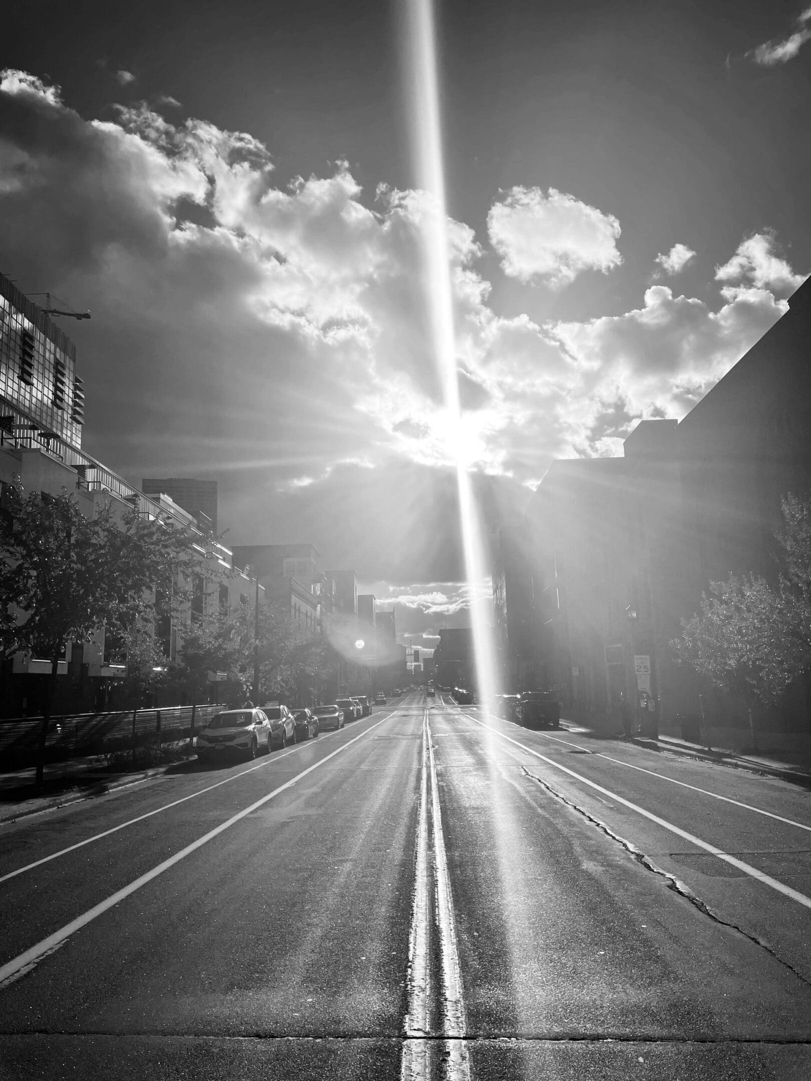 Black and white photo of an empty city street with sun rays breaking through clouds.