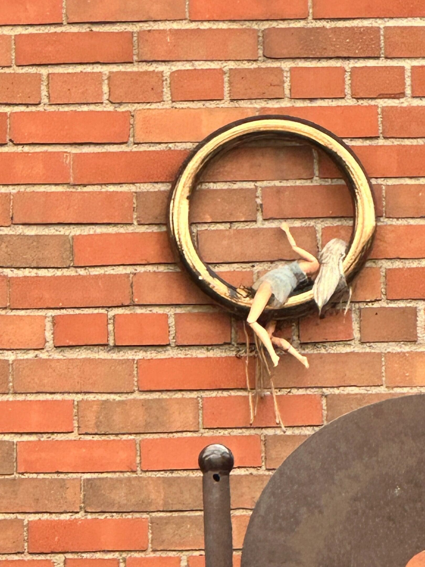 A doll hanging on a circular object against a brick wall.