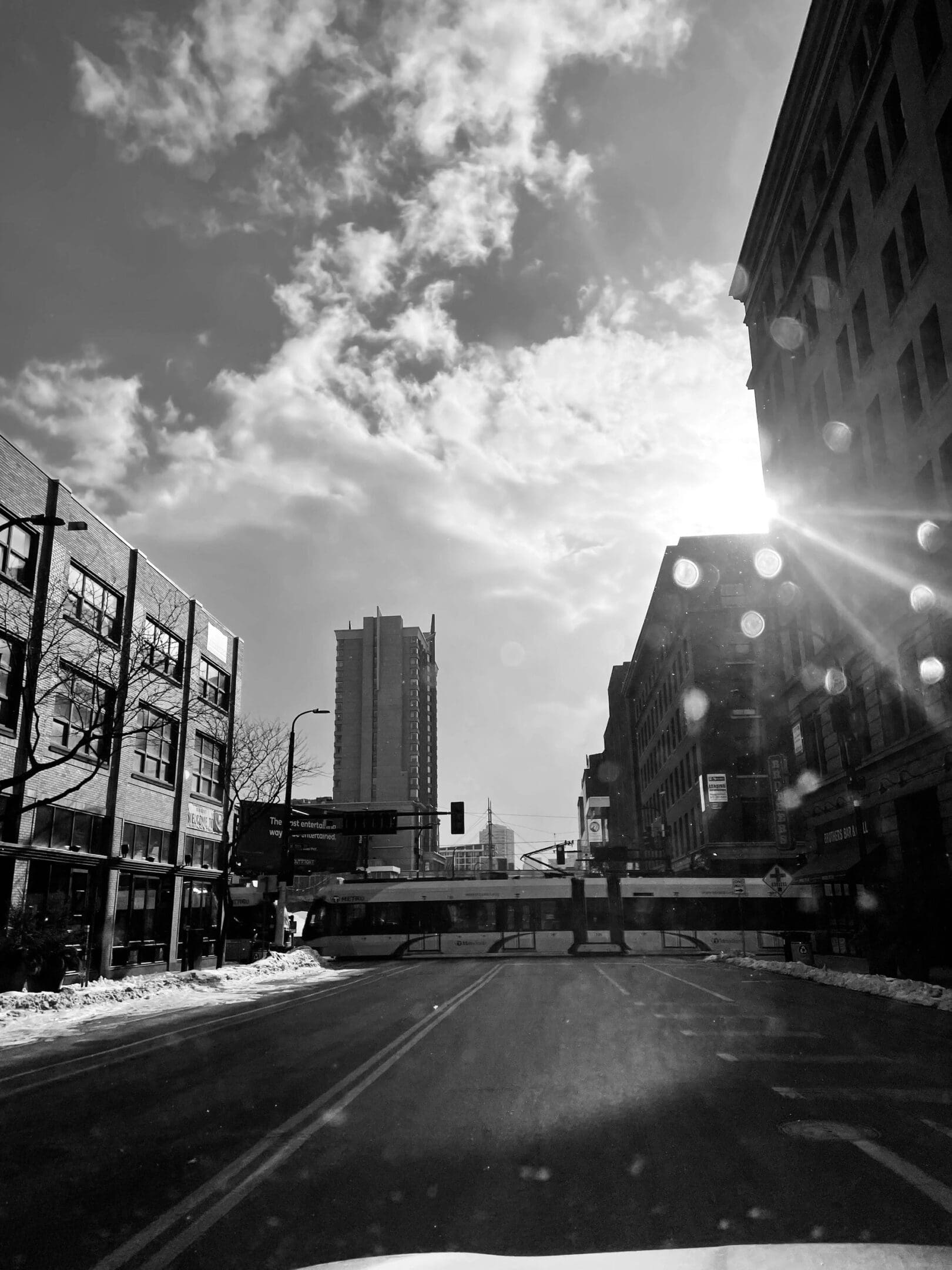 Black and white urban street scene with a light rail train crossing and sunlight.