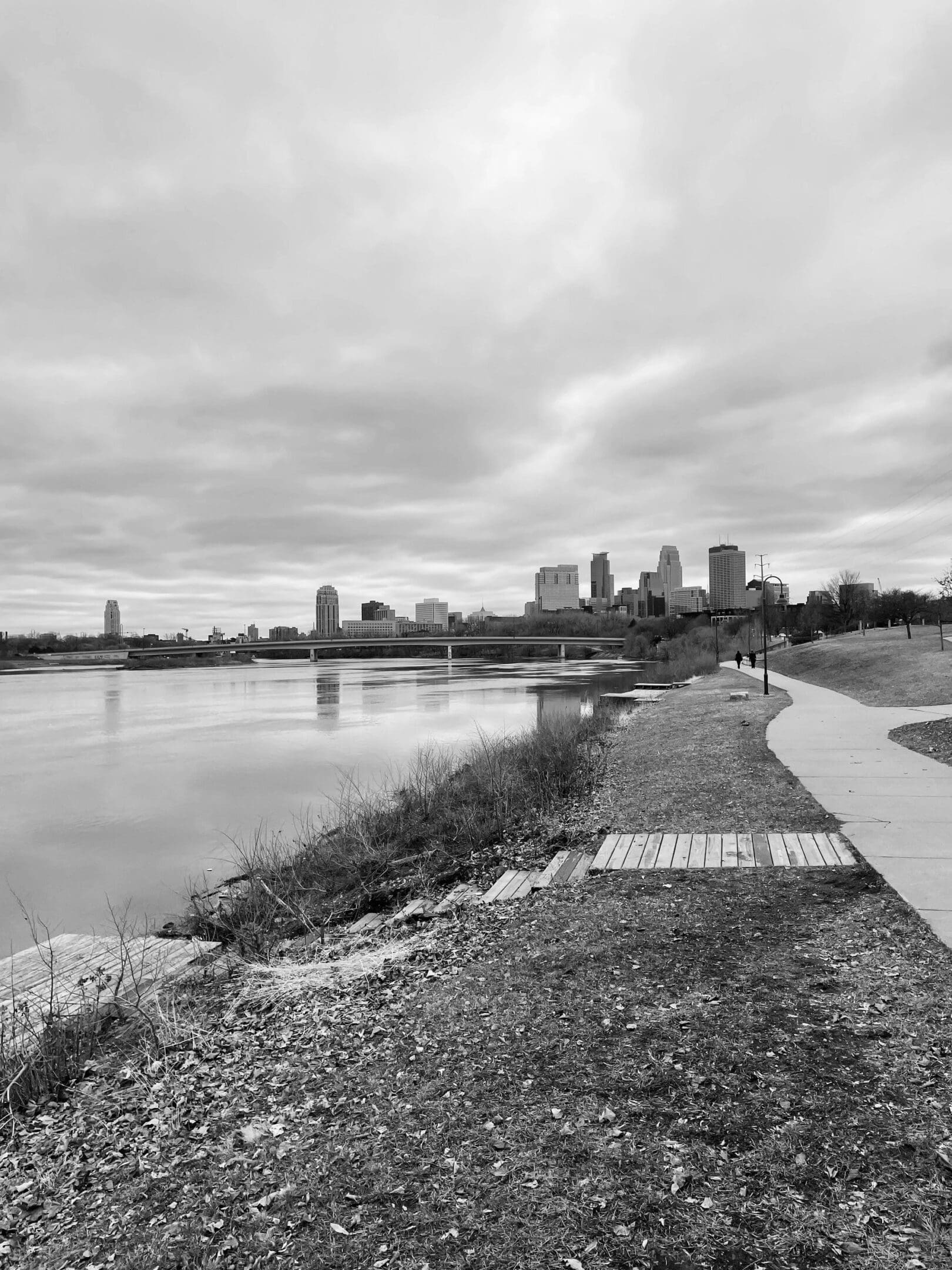 Black and white cityscape with river and walking path under cloudy sky.